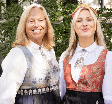 En ung og en eldre blond dame. Står foran en grønn busk ikledd bunad.